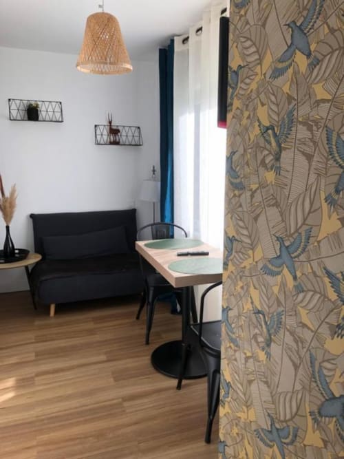 Studio Supérieur Cosyade à Bourbourg avec canapé noir, table à manger, luminaire suspendu tressé et papier peint botanique