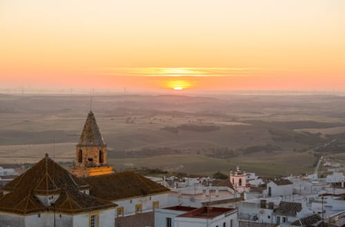 Puesta de sol sobre Medina Sidonia con la torre de la iglesia histórica y las casas blancas del pueblo dominando el paisaje rural.