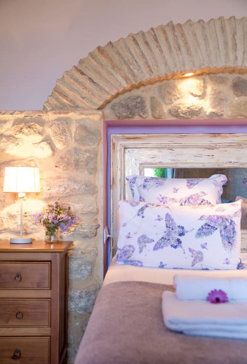 Acogedor dormitorio en apartamento de Medina Sidonia con muros de piedra, almohadas florales púrpura, mesilla de madera e iluminación ambiental