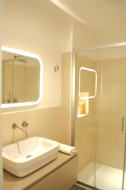 Bagno moderno con specchio illuminato, lavabo bianco a forma di vasca e doccia in vetro presso la boutique Le Dimore Del Duca Roma