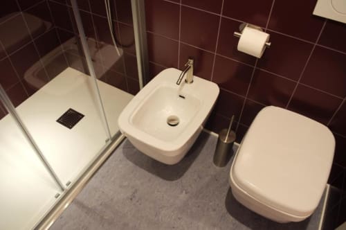 Bagno moderno presso Le Dimore Del Duca Roma con bidet bianco, wc, lavabo e doccia con box in vetro e piastrelle scure.