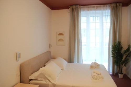 Camera luminosa del Le Dimore Del Duca Roma con letto, tende beige, porte finestre, pianta e illuminazione calda