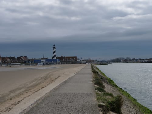 Coastal view of Bourbourg with sandy beach, striped lighthouse, and waterfront buildings under cloudy sky