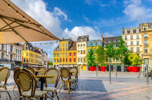 Sunny European plaza with café seating, colorful buildings, and red planters in Bourbourg, France