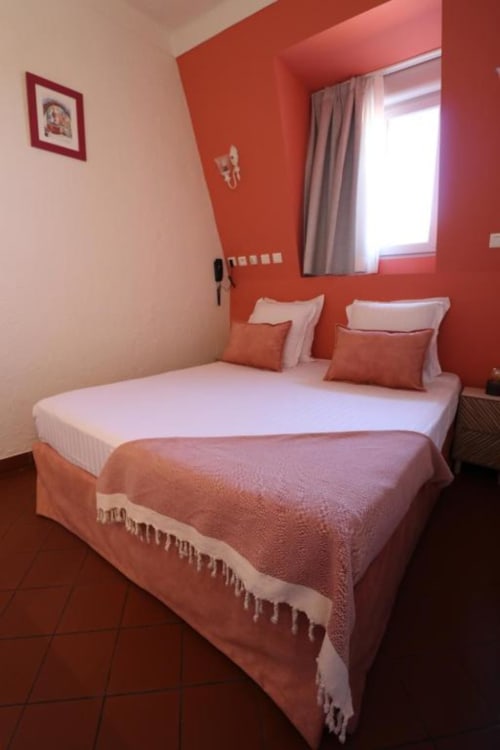 Chambre moderne à l'Hôtel Villa Roseraie à Vence avec murs accentués de rouge, lit blanc, coussins orange et fenêtre aux rideaux