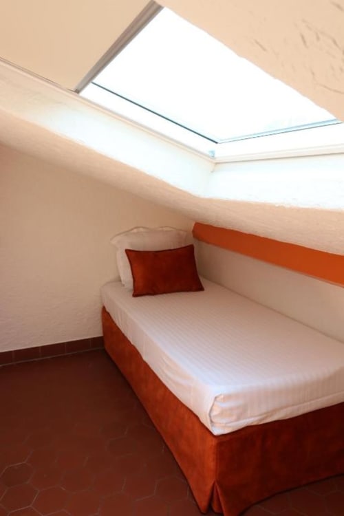 Chambre mansardée moderne à l'Hôtel Villa Roseraie à Vence avec lucarne, literie blanche, coussin rouge et parquet foncé.
