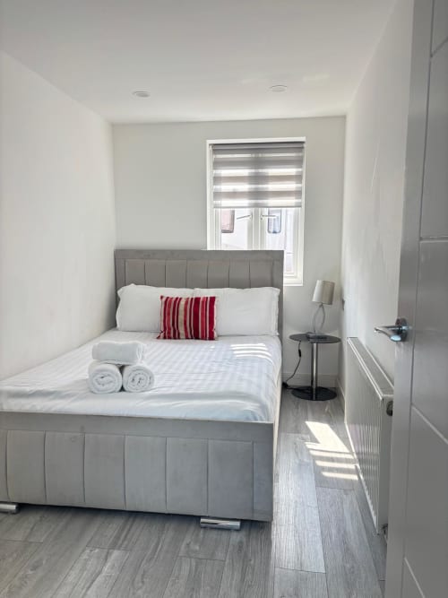 Modern bedroom with gray upholstered bed, white linens, red striped pillow, window with blinds, and clean minimalist décor at