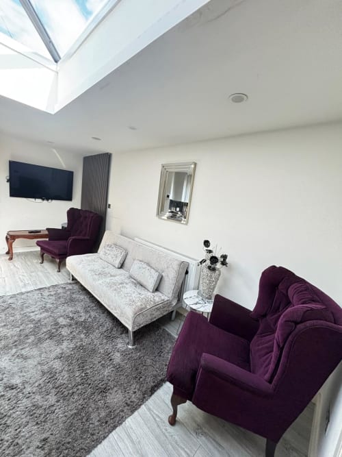Modern living room with purple velvet chairs, white sofa, TV, skylight, and garden view at England Furnished Homes in High