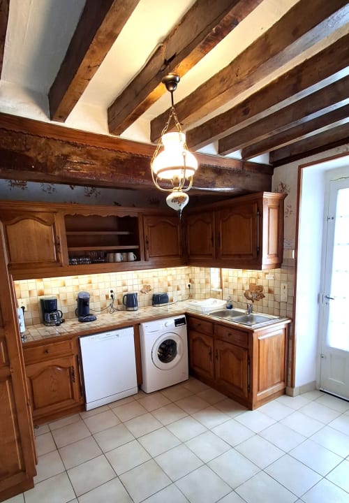 Rustic kitchen with exposed wooden beams, tiled backsplash, wooden cabinetry, washer/dryer, and pendant light at Les Gîtes de