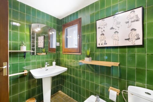Baño de azulejos verde brillante con lavamano pedestal, espejos de madera, plantas en maceta y arte mural lúdico en Masía Rural Ca l'Eloi