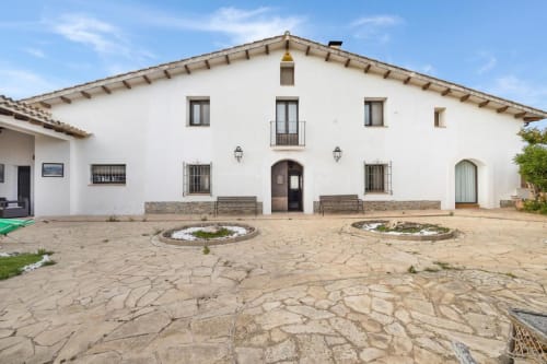 Masía Rural Ca l'Eloi Janet, patio tradicional en Barcelona con casa de piedra blanca, terraza pavimentada y elementos vegetales