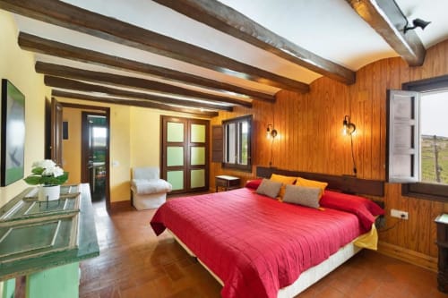 Spacious bedroom at Masia Rural Ca l'Eloi Janet near Barcelona with red bedding, wooden beams, mountain views, and warm