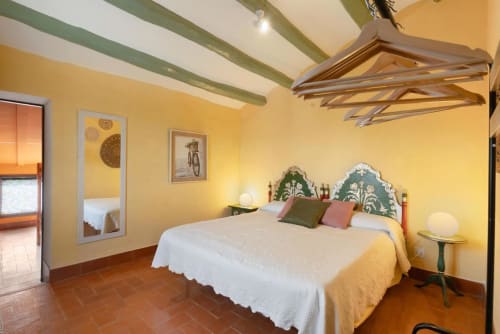 Dormitorio amarillo brillante con viga verde en el techo, cabecera ornamentada y pisos de terracota rústica en Masia Rural Ca l'Eloi Janet.