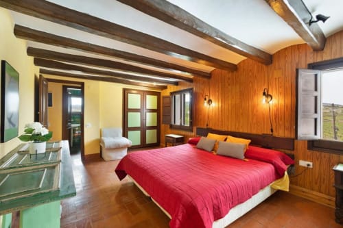 Amplio dormitorio en Masia Rural Ca l'Eloi Janet cerca de Barcelona con ropa de cama roja, vigas de madera y vistas al campo