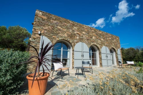 Villa Camille, Centuri : bâtisse en pierre aux fenêtres cintrées, terrasse avec mobilier, plante en pot sous ciel dégagé