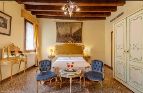 Camera da letto veneziana lussuosa con testiera dorata ornamentale, sedie blu e travi in legno a vista al Palazzo San Firmino, Venezia