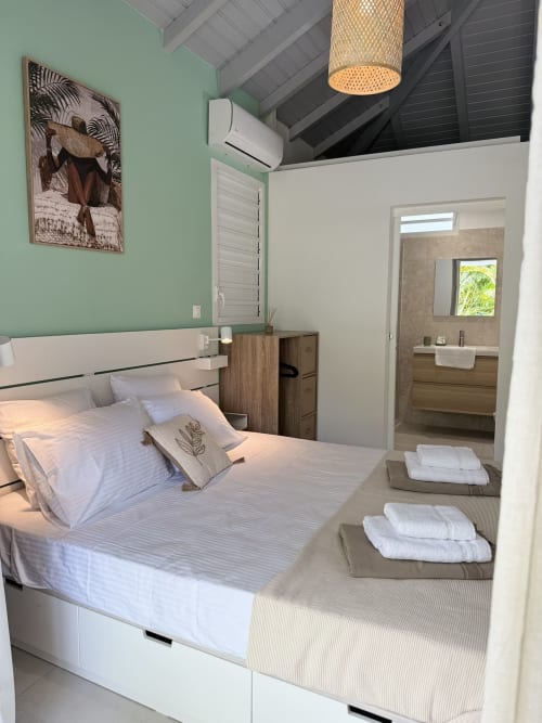 Chambre moderne du Domaine de la Glacière aux murs vert menthe, lit blanc, table de nuit en bois et salle de bain attenante à Petit