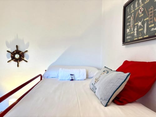 Dormitorio con temática náutica en villa Vila Natercia, Baiona, con reloj rueda de barco, ropa de cama blanca, almohadas rojas y vistas al mar