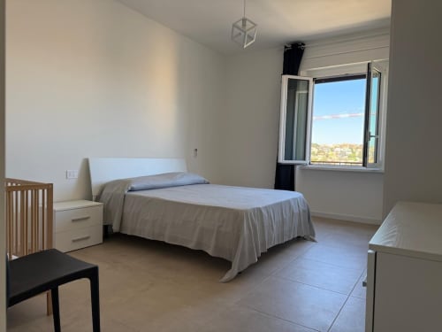 Camera da letto luminosa in appartamento romano con letto, finestra con vista sulla città, arredamento moderno e pavimento in ceramica