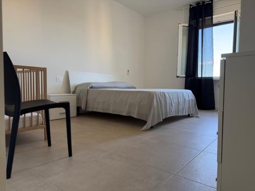 Camera da letto luminosa in appartamento romano con letto, sedia in legno, pavimento in piastrelle e tende nere che incorniciano la finestra con vista sulla città