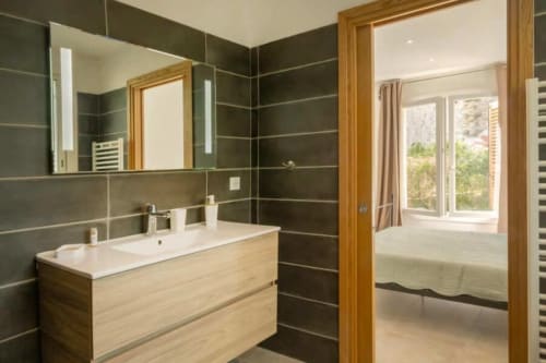 Salle de bains moderne à la Villa Odette, La Conciergerie du Cap Corse, avec murs en carrelage foncé, meuble vasque en bois et vue sur la chambre
