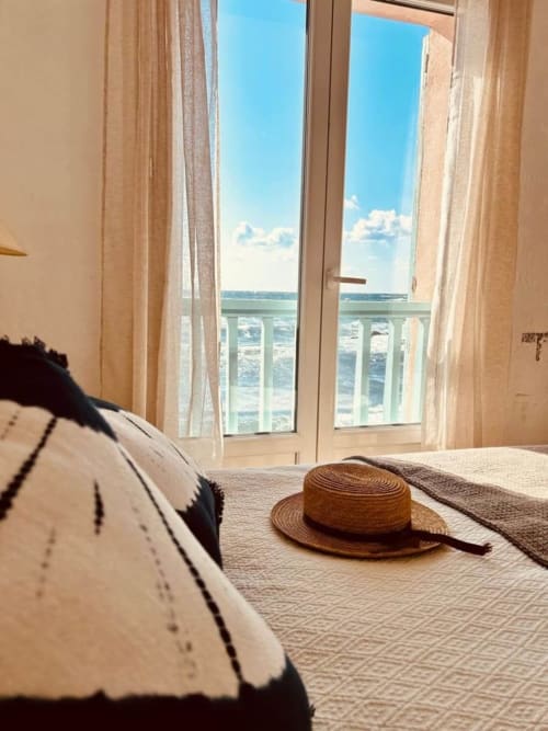 Chambre de villa bord de mer à Centuri avec portes-fenêtres ouvertes, vue sur la mer, chapeau de paille sur le lit et ambiance méditerranéenne lumineuse