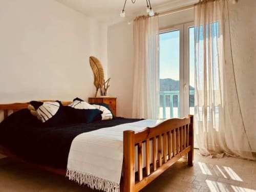 Chambre lumineuse avec lit en bois, vue sur la mer et les montagnes à travers les portes vitrées de la Villa Casapalmea, Centuri, Corse