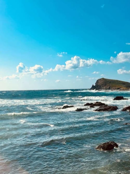 Mer turquoise aux vagues blanches et rochers sombres de la plage de Centuri, Cap Corse, avec promontoire verdoyant sous ciel bleu