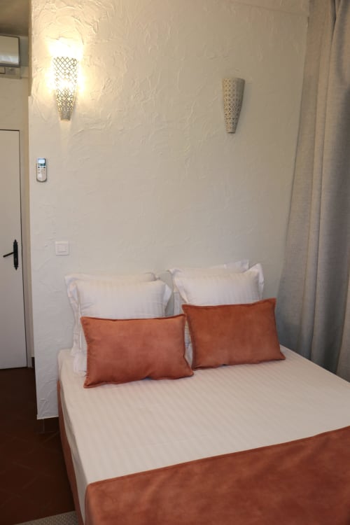 Chambre moderne à l'Hôtel Villa Roseraie avec lit blanc, oreillers couleur rouille, appliques murales, climatisation et rideaux beige