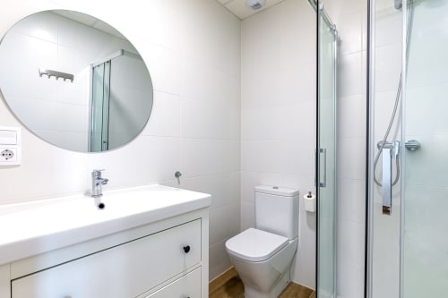 Modern bathroom at Residencia Demar with white vanity, round mirror, toilet, and walk-in shower with glass enclosure