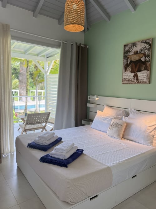 Chambre lumineuse au Domaine de la Glacière avec murs vert menthe, lit blanc, luminaire suspendu tressé et vue sur la piscine