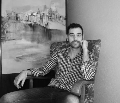 Hombre con camisa de cuadros sentado en una silla del hotel MoneoHeritage en Salamanca con una obra de arte del castillo detrás