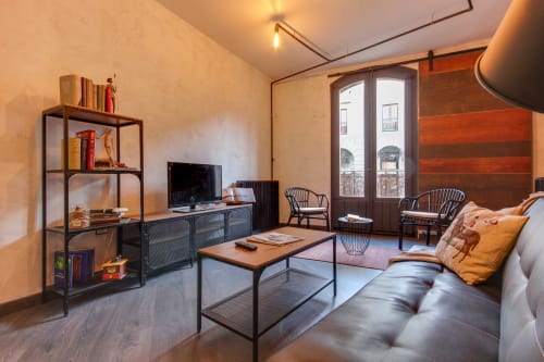 Sala de estar de loft industrial en hotel Salamanca con sofá, TV, escritorio, estantería metálica y vistas de la ciudad desde el balcón