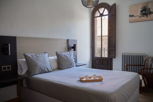 Luminosa habitación con ropa de cama blanca, almohadas grises y puertas de madera que abren a balcón con vistas a la ciudad en hotel de Salamanca