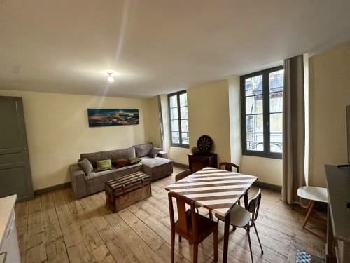 Odnowiony apartament T2 w Lourdes z stołem jadalnym, szarą sofą, drewnianymi podłogami i balkonem z widokiem na ogród
