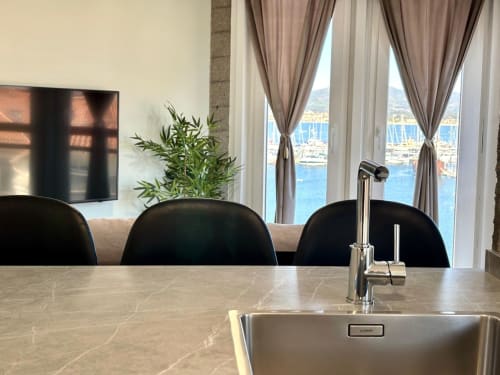 Cocina moderna con vista al mar en apartamento de Baiona. Encimera de mármol, fregadero de acero inoxidable, sillas negras, océano y barcos visibles