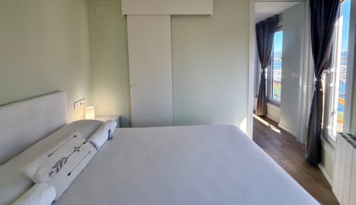Dormitorio moderno con vistas al mar en Holibai Miradoiro, Baiona. Cama doble, ropa de cama blanca, ventanas de balcón con vistas al agua.
