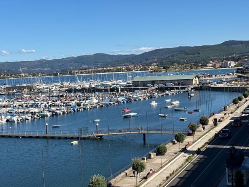 Marina de Baiona con veleros amarrados, paseo marítimo y colinas boscosas bajo cielo azul