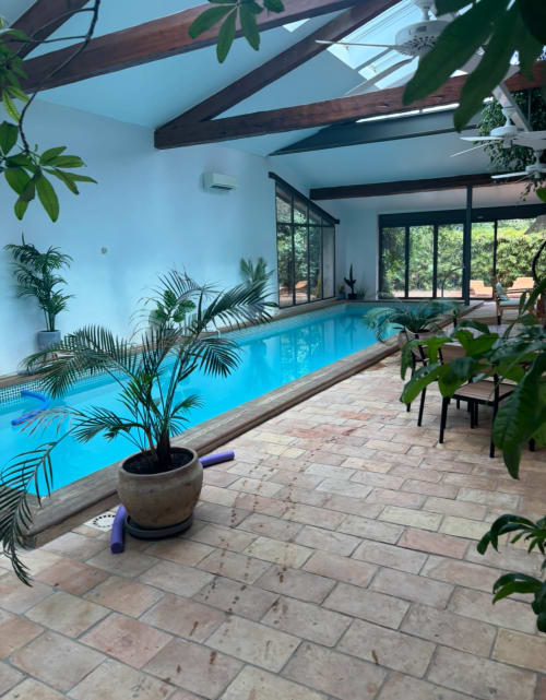 Piscine intérieure de la villa La Conciergerie De Franck à Boissieres avec plantes tropicales, poutres en bois et grandes fenêtres