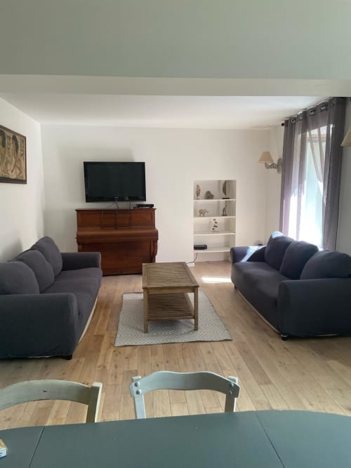 Modern living room with two dark sofas, wooden coffee table, TV on piano, and white shelving in La Conciergerie De Franck