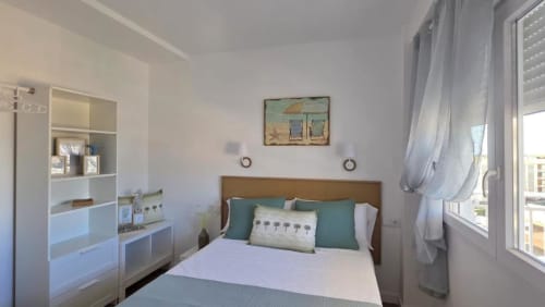 Dormitorio luminoso con cama de madera, almohadas turquesas, tocador blanco y vistas al océano desde el balcón del apartamento en Benicàssim