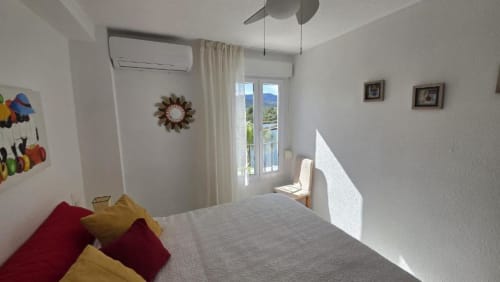 Dormitorio luminoso en el apartamento Regata, Benicàssim, con cama, almohadas coloridas, puertas a balcón, ventilador y vistas al lago
