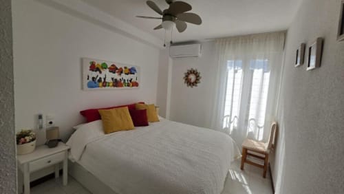 Dormitorio luminoso en el Apartamento Regata, Benicàssim con cama blanca, almohadas coloridas, ventilador de techo y puertas balconeras al lago