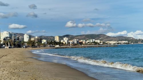 Playa de Benicàssim con arena blanca, olas del Mediterráneo, edificios de apartamentos frente al mar y montañas al fondo