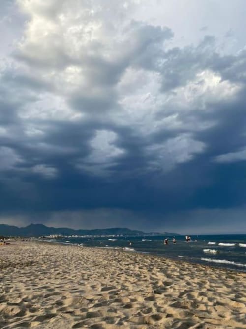 Playa arenosa en Benicàssim con bañistas y olas turquesas, cielo nublado dramático y montañas