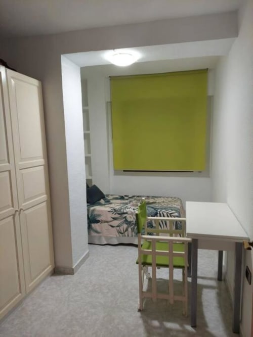 Interior de apartamento luminoso con mesa de comedor, cama con ropa de cama estampada, persiana enrollable verde lima, armario blanco y baldosas