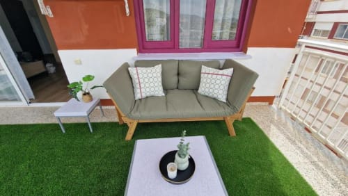 Sofá verde oliva con patas de madera y cojines estampados en terraza de césped artificial, con planta macetada y mesita lateral