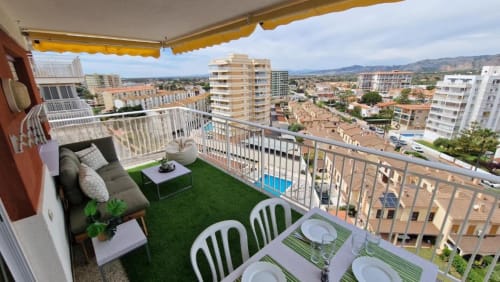 Terraza de apartamento al atardecer en Benicàssim con césped artificial, mesa de comedor, zona de descanso y vistas panorámicas de la localidad costera