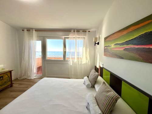 Dormitorio luminoso con vistas al mar en  Holibai Baiona, con ropa de cama blanca, detalles verde lima y balcón frente al océano