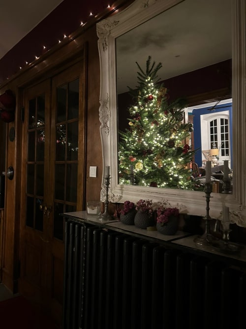 Élégante entrance de la maison d’hôtes Le Clos d'Azel avec sapin de Noël illuminé se reflétant dans un miroir ancien, radiateur en fonte anthracite et ambiance festive d’un Noël traditionnel en Alsace.